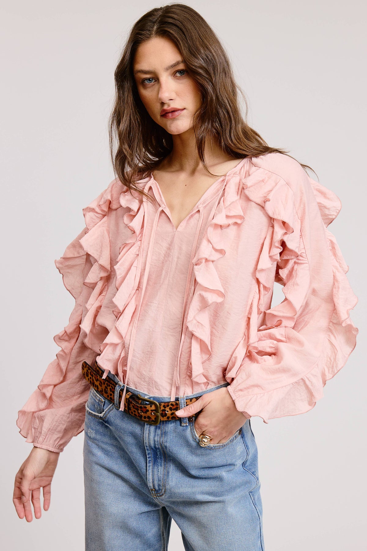 A woman wearing a light pink long-sleeve blouse with dramatic layered ruffles and a V-neckline, paired with blue jeans and a brown patterned belt, against a light gray studio background.