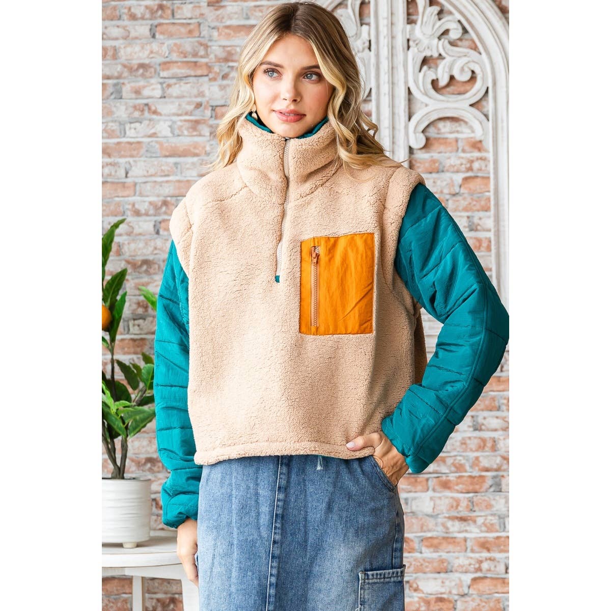 Woman wearing a beige vest with an orange pocket and teal sleeves against a brick wall.