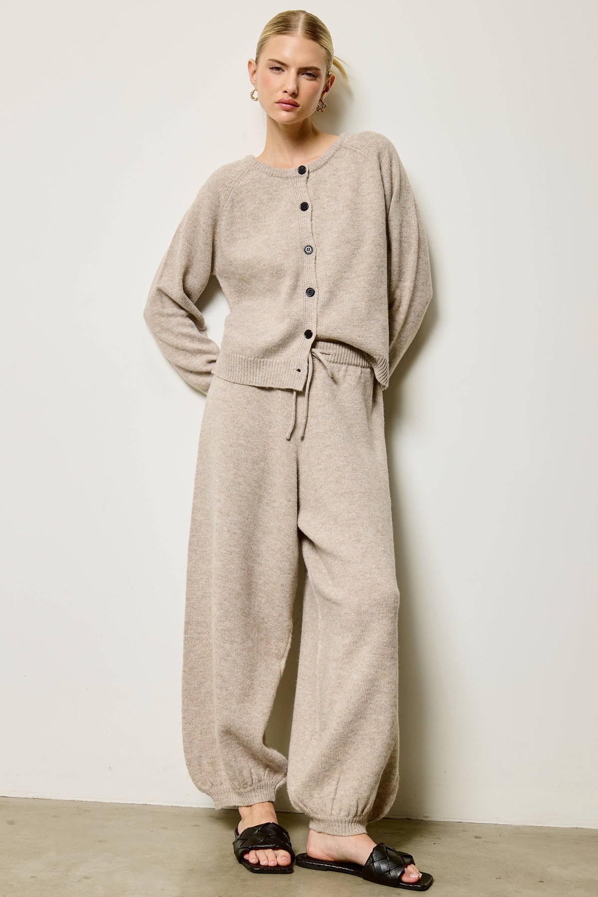 A woman modeling a light beige knit loungewear set consisting of a long-sleeve button-front top and matching wide-leg pants, paired with black slide sandals against a plain off-white wall.
