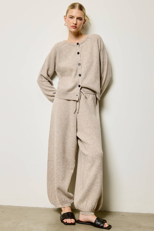 A woman modeling a light beige knit loungewear set consisting of a long-sleeve button-front top and matching wide-leg pants, paired with black slide sandals against a plain off-white wall.