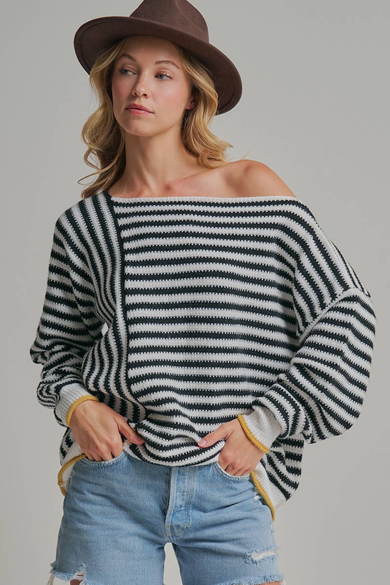 A woman wearing a black and white horizontally striped off-the-shoulder knit sweater, light blue distressed denim shorts, and a brown wide-brim hat, posed against a solid light gray studio background.