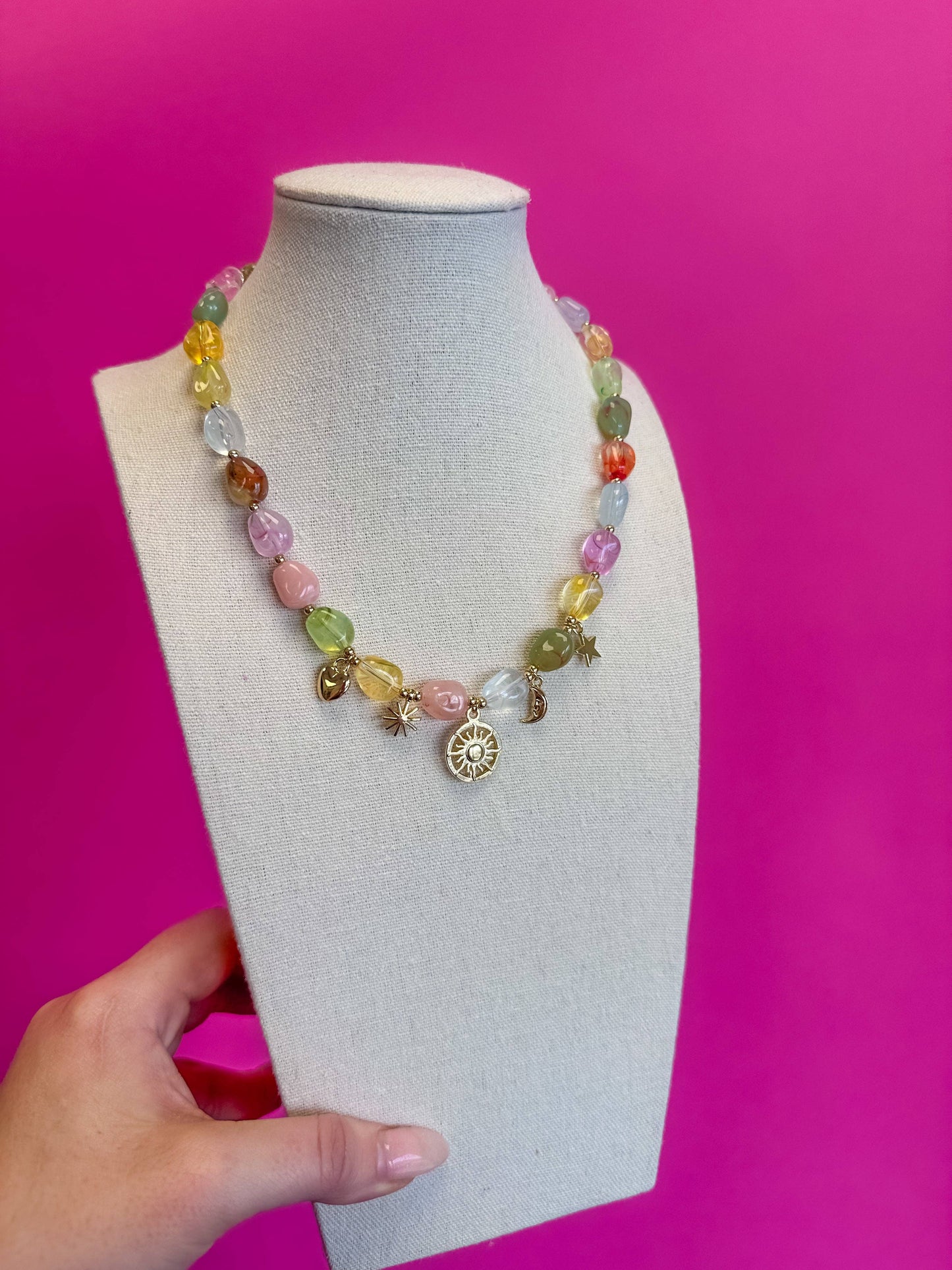 A multicolor beaded necklace with gold-toned charms, displayed on a light gray fabric jewelry bust held by a hand against a bright pink background.