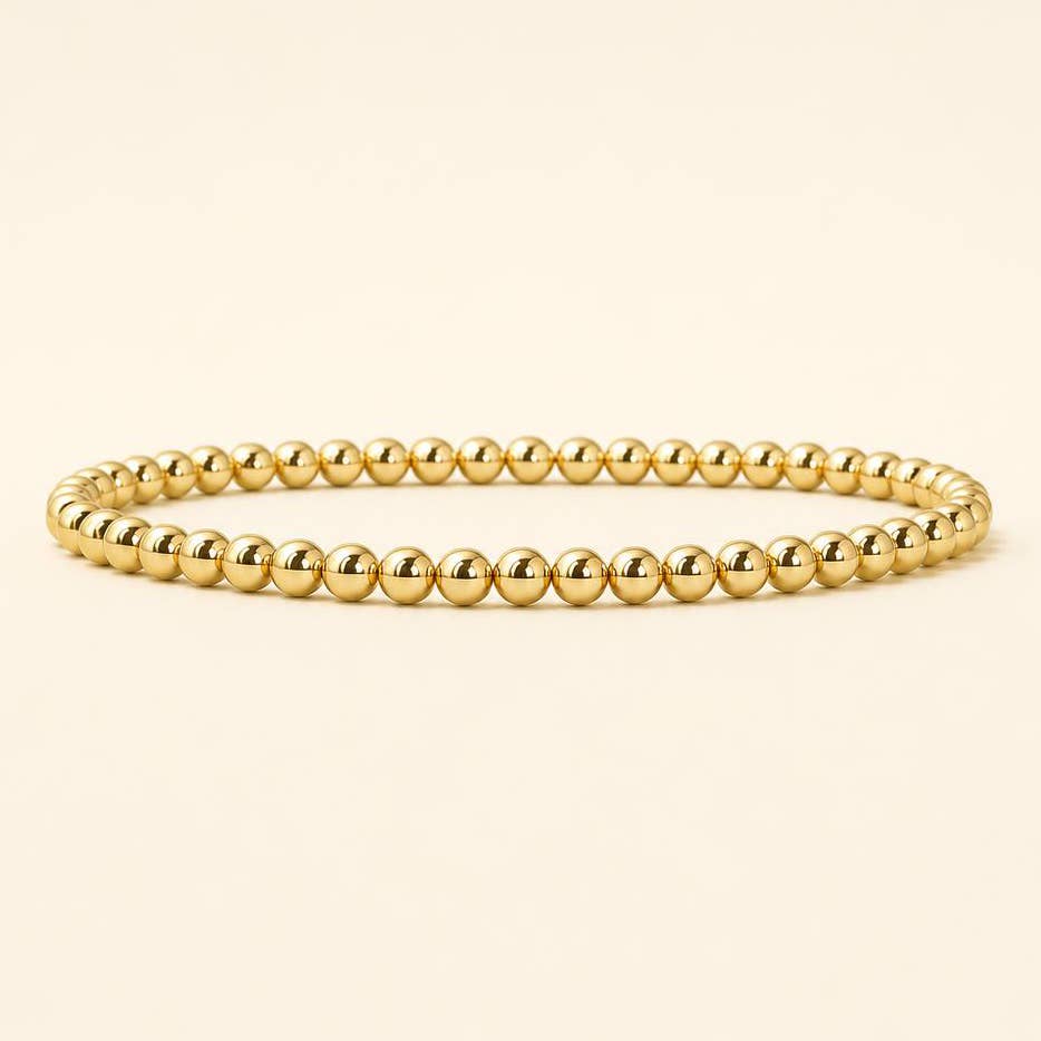 A gold beaded stretch bracelet with polished, rounded spherical beads, displayed on a clean, solid cream background.