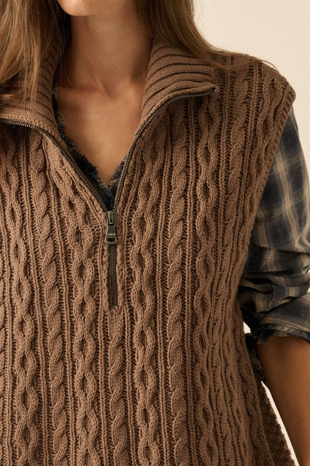 Close-up of a person wearing a brown cable knit vest with a neutral background