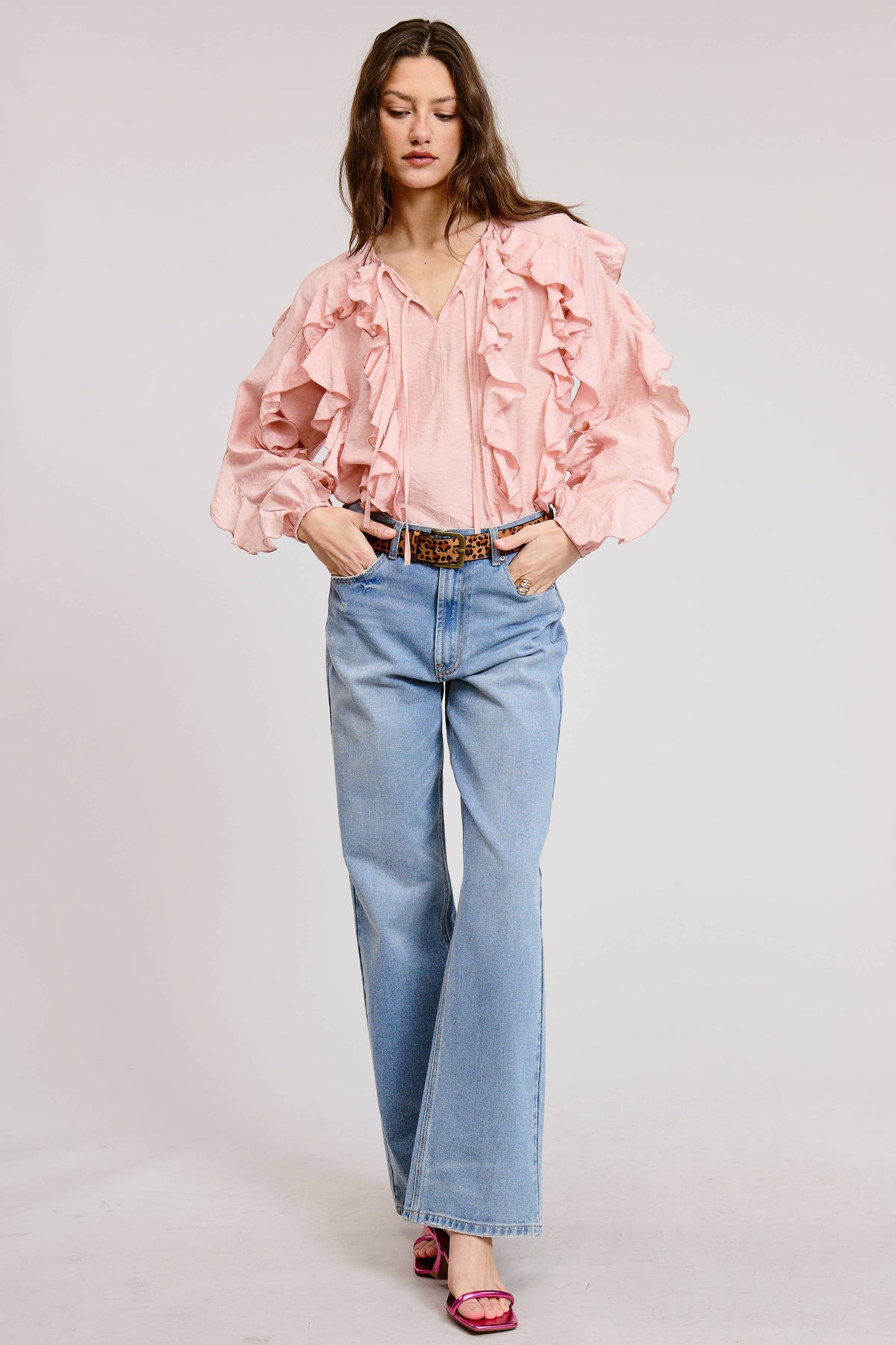 A woman modeling a light pink ruffled blouse with voluminous sleeves, paired with light blue wide-leg jeans and pink sandals, against a plain light gray studio background.