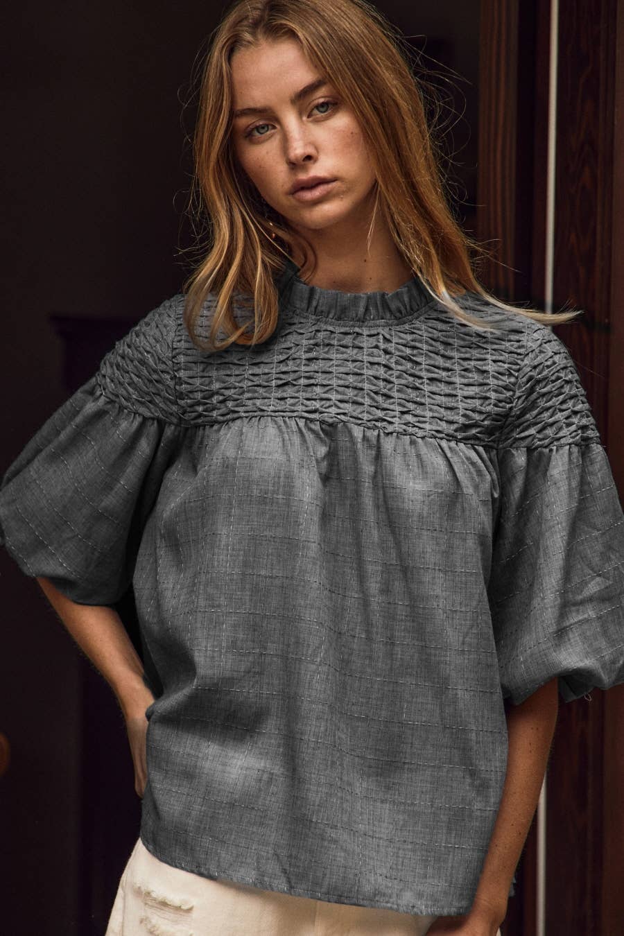 A woman wearing a dark gray smocked blouse with short puff sleeves and a gathered neckline, paired with light beige pants, set against a dark background.