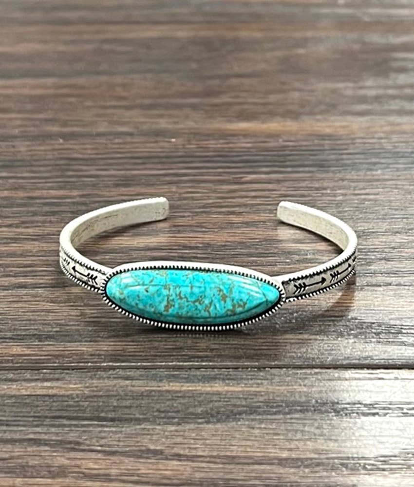 A silver cuff bracelet with a central oval turquoise stone, resting on a dark wood surface.