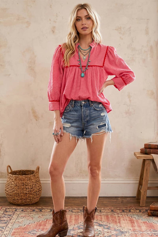 A woman modeling a pink long-sleeve blouse with ruffled trim, paired with distressed denim shorts and brown cowboy boots, standing in a rustic room with a textured beige wall, woven basket, and wooden stool.