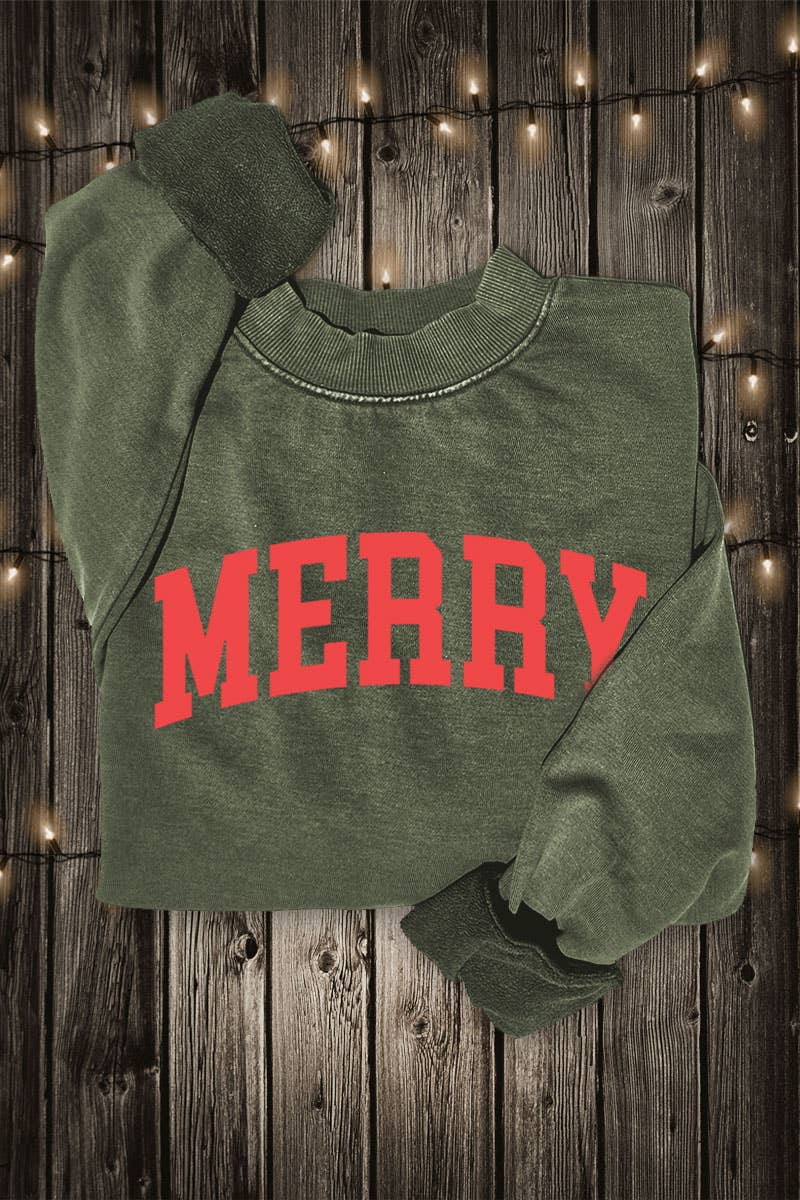 An olive green crewneck sweatshirt with 'MERRY' printed in red arched block letters across the chest, laid flat on a dark wood plank background with warm string lights.