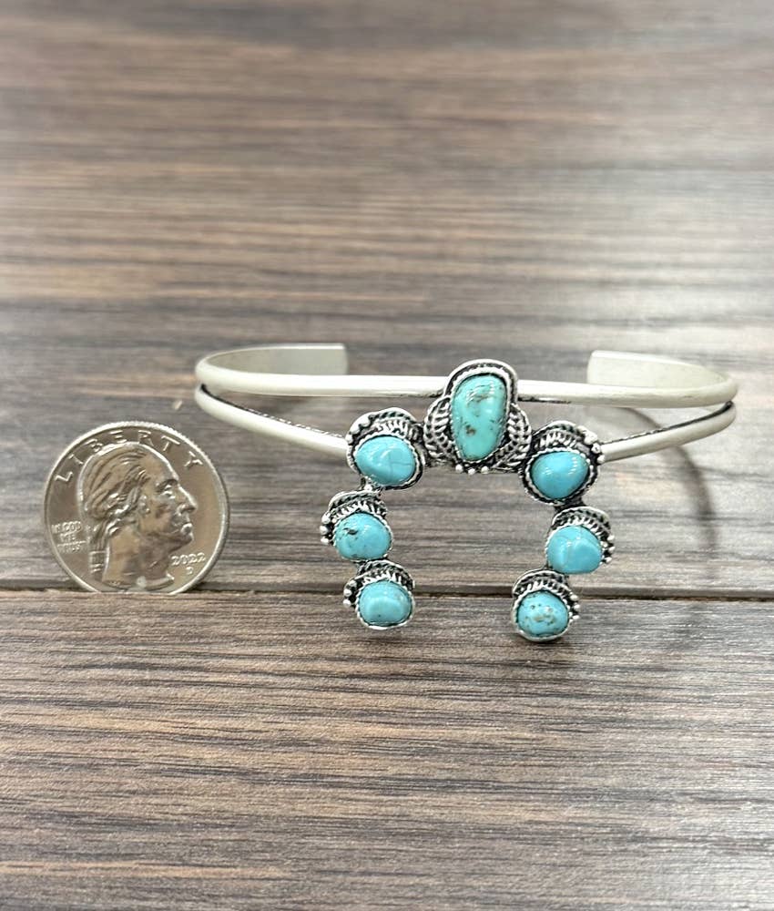 A sterling silver cuff bracelet with six turquoise stones, displayed on a wooden surface next to a US quarter for scale.