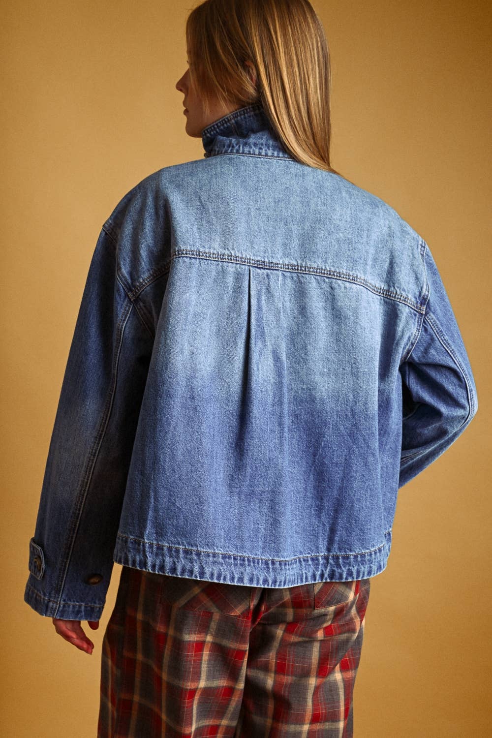 Person wearing a blue denim jacket and plaid pants against a beige background
