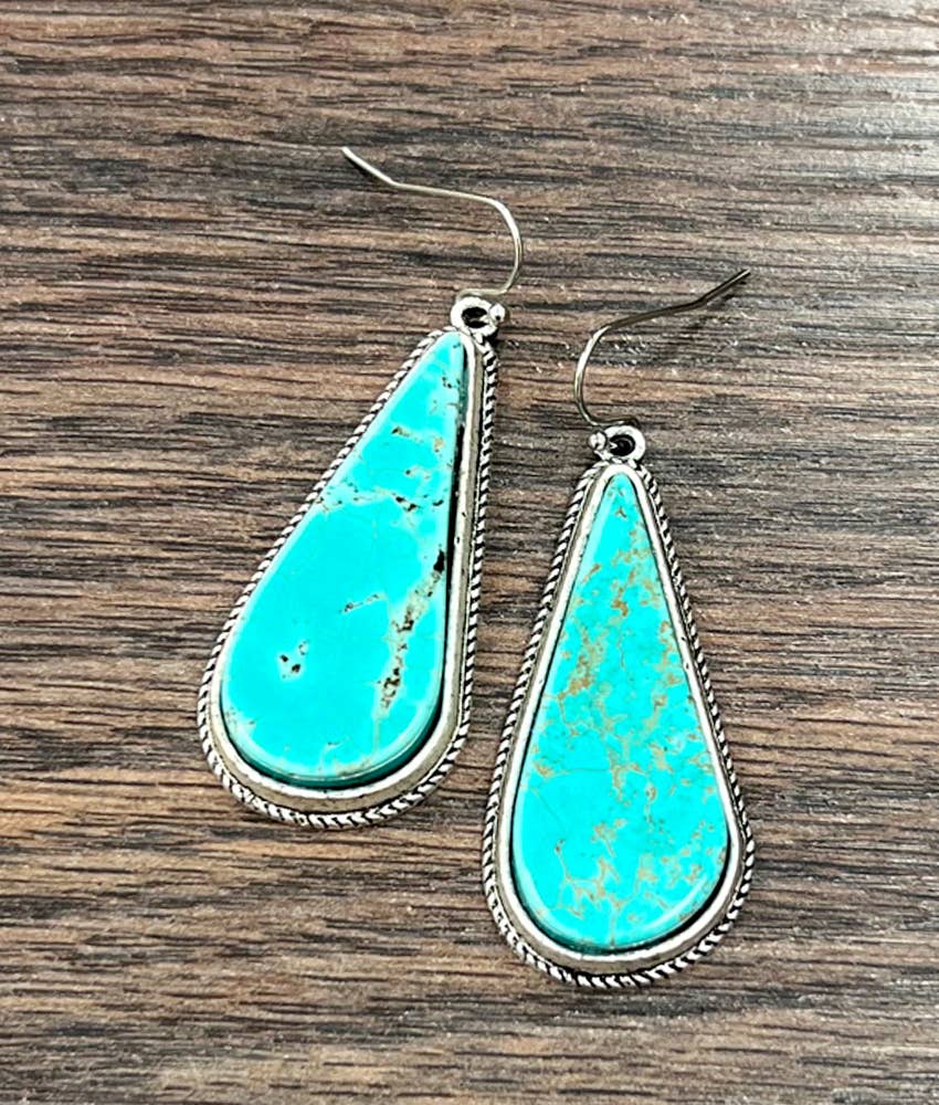 A pair of teardrop-shaped turquoise drop earrings with silver-toned twisted border settings and fishhook wires, displayed on a dark wood background.