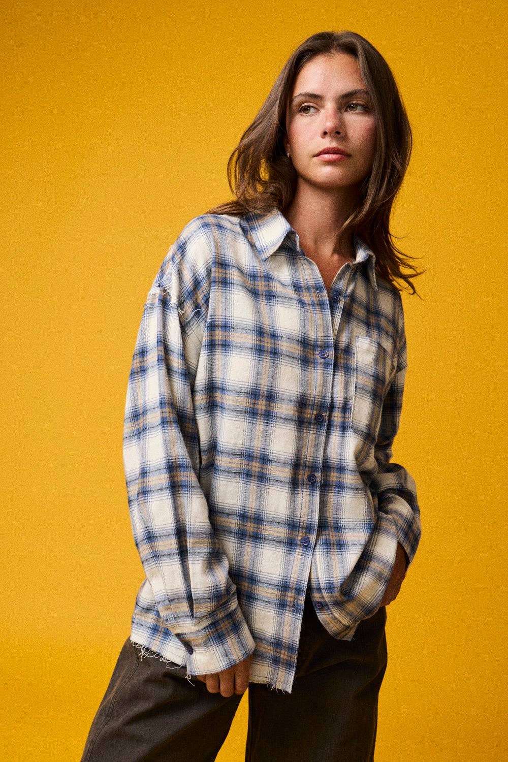 A woman modeling a long-sleeve plaid button-up shirt in white, blue, and gray, paired with dark brown pants, set against a solid mustard yellow background.