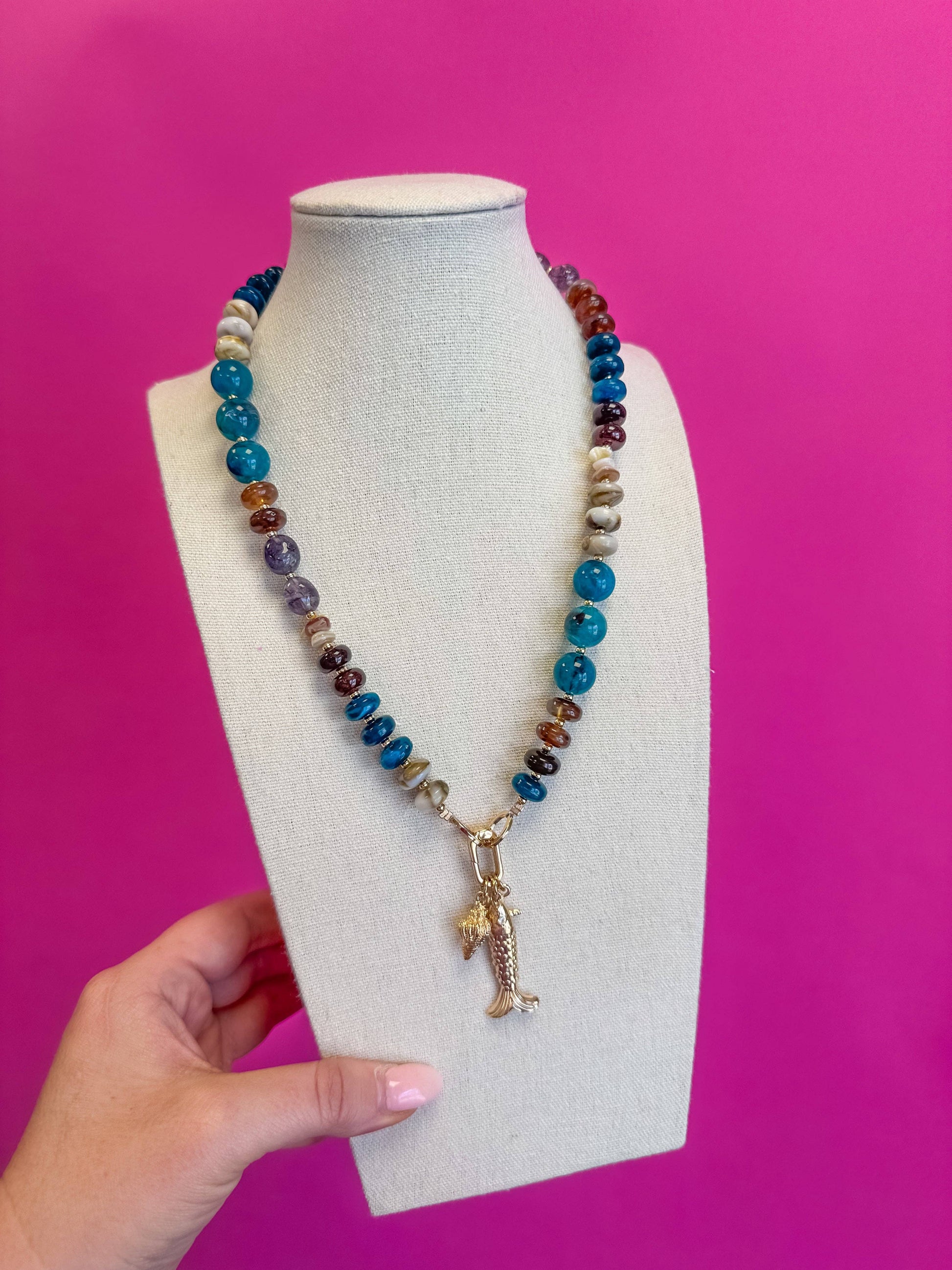 A beaded necklace with turquoise-blue, brown, and white beads, featuring a gold-toned mermaid charm, displayed on a cream-colored jewelry bust with a hand holding the base against a bright pink background.